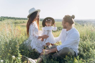 Happy young family spending  time together in nature, beautiful woman in white guipure dress with husband and their daughter sitting in the field