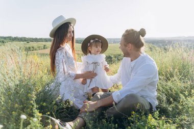 Happy young family spending  time together in nature, beautiful woman in white guipure dress with husband and their daughter sitting in the field