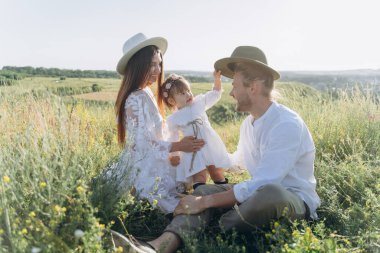 Happy young family spending  time together in nature, beautiful woman in white guipure dress with husband and their daughter sitting in the field