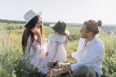 Happy young family spending  time together in nature, beautiful woman in white guipure dress with husband and their daughter sitting in the field
