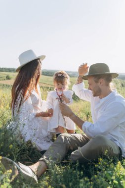 Happy young family spending  time together in nature, beautiful woman in white guipure dress with husband and their daughter sitting in the field