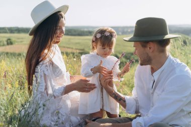 Happy young family spending  time together in nature, beautiful woman in white guipure dress with husband and their daughter sitting in the field