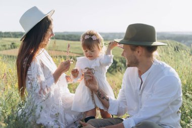 Happy young family spending  time together in nature, beautiful woman in white guipure dress with husband and their daughter sitting in the field