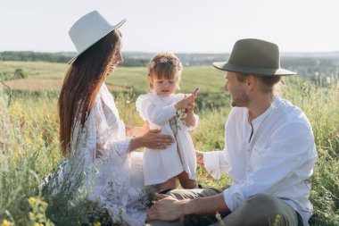 Happy young family spending  time together in nature, beautiful woman in white guipure dress with husband and their daughter sitting in the field