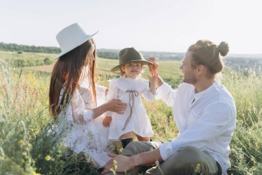 Happy young family spending  time together in nature, beautiful woman in white guipure dress with husband and their daughter sitting in the field