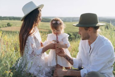 Happy young family spending  time together in nature, beautiful woman in white guipure dress with husband and their daughter sitting in the field
