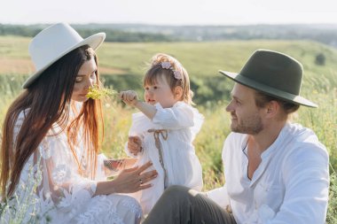 Happy young family spending  time together in nature, beautiful woman in white guipure dress with husband and their daughter sitting in the field