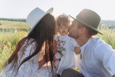 Happy young family spending  time together in nature, beautiful woman in white guipure dress with husband kissing their daughter 