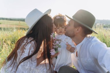 Happy young family spending  time together in nature, beautiful woman in white guipure dress with husband kissing their daughter 