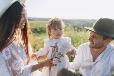 Happy young family spending  time together in nature, beautiful woman in white guipure dress with husband and their daughter sitting in the field