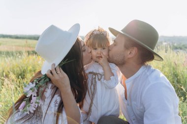 Happy young family spending  time together in nature, beautiful woman in white guipure dress with husband kissing their daughter 