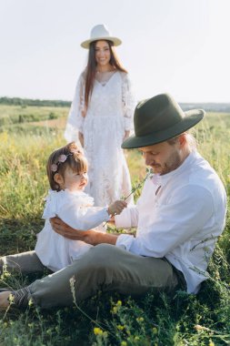 Happy young family spending  time together in nature, beautiful woman in white guipure dress with husband and their daughter sitting in the field
