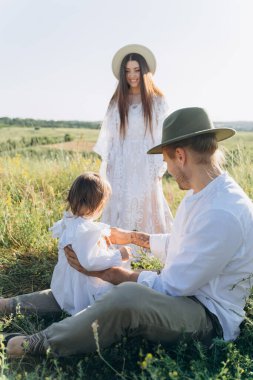 Happy young family spending  time together in nature, beautiful woman in white guipure dress with husband and their daughter sitting in the field