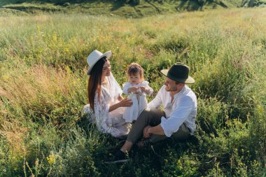 Happy young family spending  time together in nature, beautiful woman in white guipure dress with husband and their daughter sitting in the field