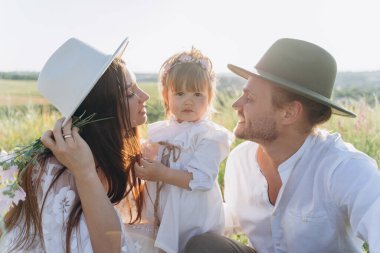 Happy young family spending  time together in nature, beautiful woman in white guipure dress with husband and their daughter sitting in the field