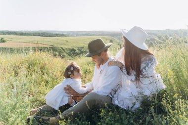 Happy young family spending  time together in nature, beautiful woman in white guipure dress with husband and their daughter sitting in the field