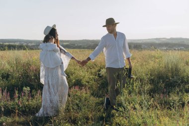 Happy young family spending  time together in nature, beautiful woman in white guipure dress holding her daughter and walking with husband in the field