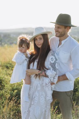 Happy young family spending  time together in nature, beautiful woman in white guipure dress holding her daughter and walking with husband in the field