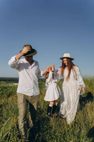 Happy young family spending  time together in nature, beautiful woman in white guipure dress with husband and their daughter walking  in the field