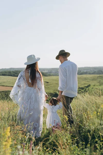 Happy young family spending  time together in nature, beautiful woman in white guipure dress with husband and their daughter walking  in the field