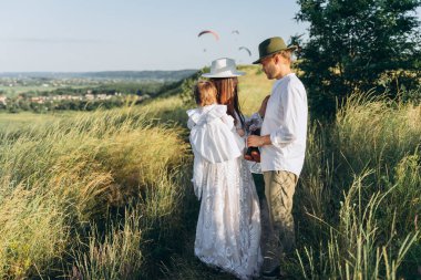 Happy young family spending  time together in nature, beautiful woman in white guipure dress holding her daughter and walking with husband in the field