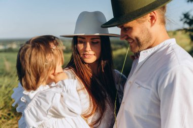 Happy young family spending  time together in nature, beautiful woman in white guipure dress holding her daughter and walking with husband in the field