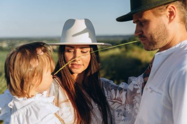 Happy young family spending  time together in nature, beautiful woman in white guipure dress holding her daughter and walking with husband in the field