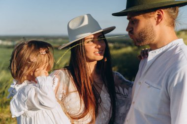 Happy young family spending  time together in nature, beautiful woman in white guipure dress holding her daughter and walking with husband in the field