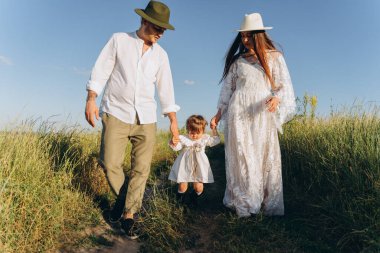 Happy young family spending  time together in nature, beautiful woman in white guipure dress with husband and their daughter walking in the field