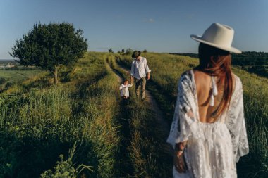Happy young family spending  time together in nature, beautiful woman in white guipure dress with husband and their daughter walking  in the field