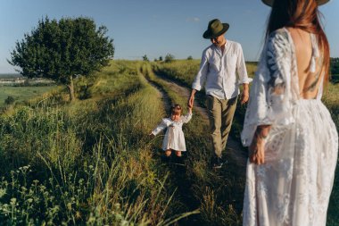 Happy young family spending  time together in nature, beautiful woman in white guipure dress with husband and their daughter walking  in the field