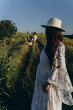 Happy young family spending  time together in nature, beautiful woman in white guipure dress with husband and their daughter walking  in the field