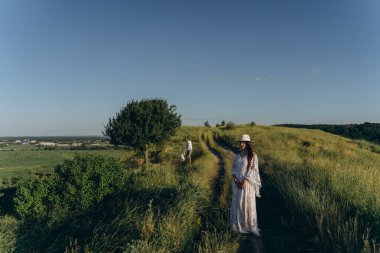 Happy young family spending  time together in nature, beautiful woman in white guipure dress with husband and their daughter walking  in the field