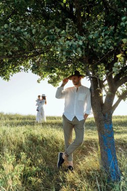 Happy young family spending  time together in nature, beautiful woman in white guipure dress holding her daughter, her husband standing by the tree in the field