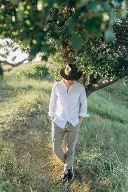 The handsome young man in a hat standing by a tree and posing at the camera