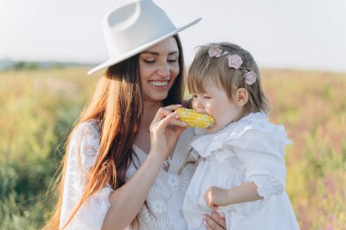 The beautiful woman in white guipure dress holding her daughter and eating corn in the field
