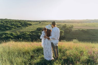 Happy young family spending  time together in nature, beautiful woman in white guipure dress holding her daughter and walking with husband in the field