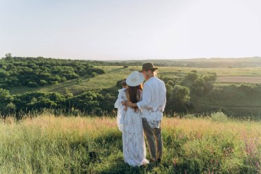 Happy young family spending  time together in nature, beautiful woman in white guipure dress holding her daughter and walking with husband in the field