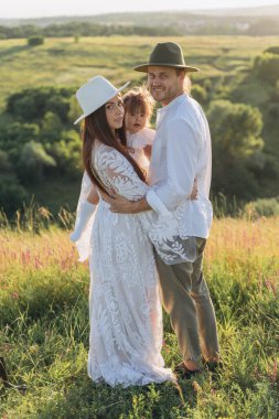Happy young family spending  time together in nature, beautiful woman in white guipure dress holding her daughter and walking with husband in the field