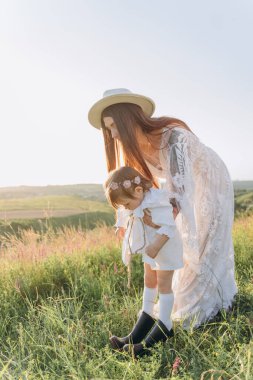 The beautiful woman in white guipure dress with her daughter sitting on the field and mother helping daughter to put on boots