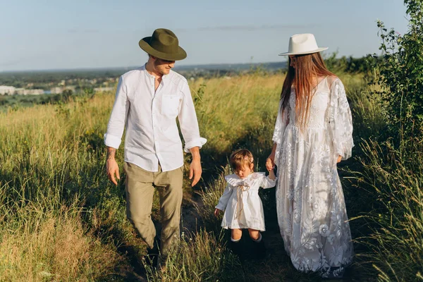 Happy young family spending  time together in nature, beautiful woman in white guipure dress with husband and their daughter walking in the field