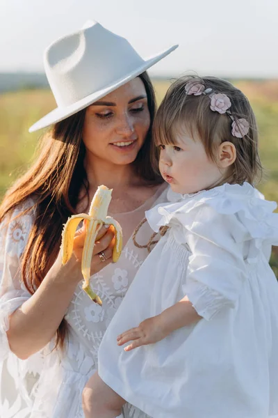 Красивая Женщина Белом Платье Guipure Держа Дочь Едят Банан Поле — стоковое фото