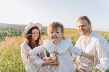 Happy young family spending  time together in nature, beautiful woman in white guipure dress with husband and their daughter sitting in the field