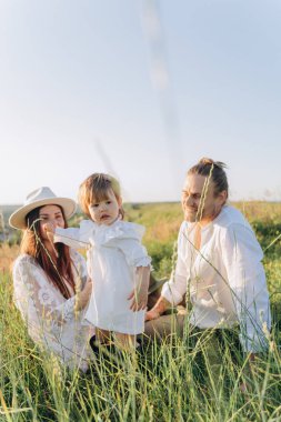 Happy young family spending  time together in nature, beautiful woman in white guipure dress with husband and their daughter sitting in the field