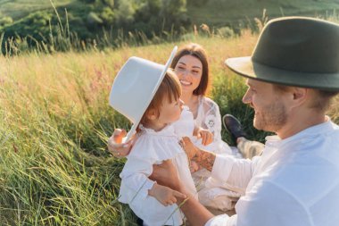 Happy young family spending  time together in nature, beautiful woman in white guipure dress with husband and their daughter sitting in the field