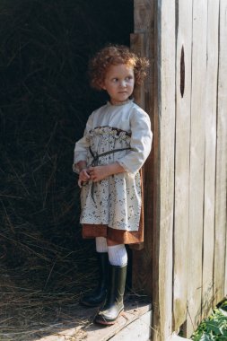 Eski moda elbiseli güzel kızıl saçlı kız çiftlikteki ahşap ahırın yanında duruyor.