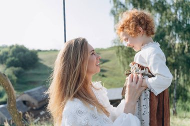 Beyaz dantelli elbiseli güzel kadın, kızıl saçlı kızıyla salıncakta oturuyor.