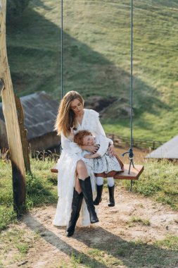 Beyaz dantelli elbiseli güzel kadın, kızıl saçlı kızıyla salıncakta oturuyor.