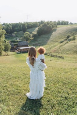 Beyaz dantel elbiseli güzel bir kadının, kızıl saçlı küçük kızıyla tarlada yürüyüşünün arka görüntüsü.  