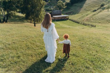 Beyaz dantel elbiseli güzel bir kadının, kızıl saçlı küçük kızıyla tarlada yürüyüşünün arka görüntüsü.  